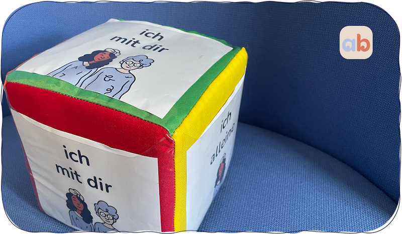 Ein weicher, farbenfroher Stoffwürfel mit deutschem Text "Ich mit dir" und Cartoon-Illustrationen von zwei Personen an den Seiten, der auf einer blauen, gepolsterten Oberfläche ruht.